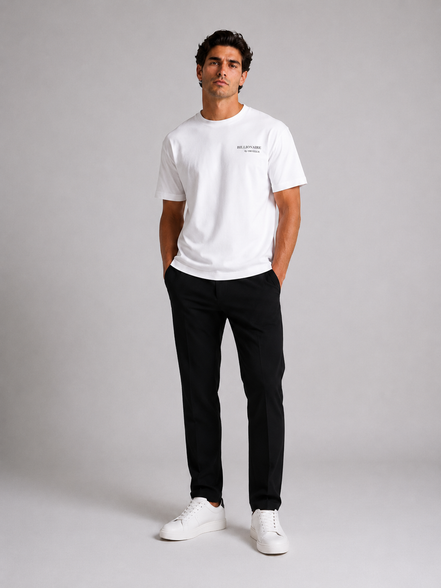 Billionaire by ORVELLA® Luxury White T-Shirt on a male model in a luxury studio photoshoot. Premium white designer T-shirt paired with black tailored trousers and white leather sneakers, showcasing modern British luxury streetwear.