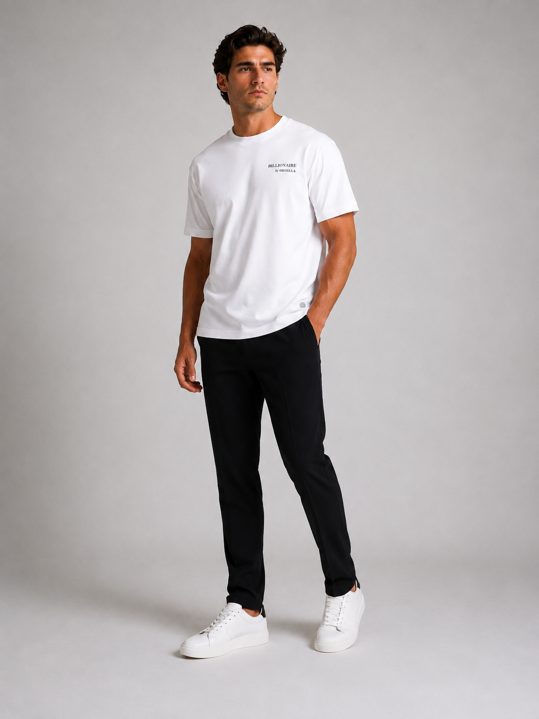 Billionaire by ORVELLA® Luxury White T-Shirt worn by a male model during a professional studio photoshoot. Premium white designer T-shirt styled with black tailored trousers and white leather sneakers, showcasing modern British luxury streetwear with a refined minimalist aesthetic.