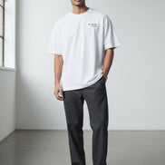 Man wearing the Billionaire by Orvella white t-shirt and dark pants standing in a minimalistic room.