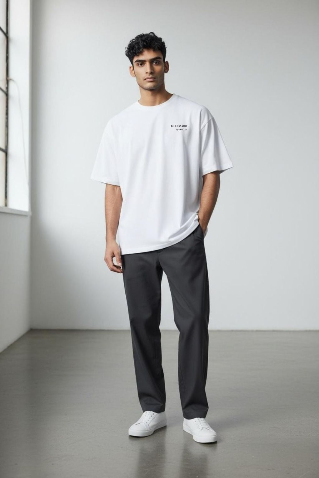 Man wearing the Billionaire by Orvella white t-shirt and dark pants standing in a minimalistic room.