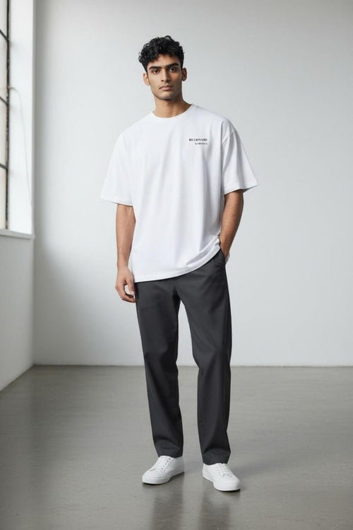 Man wearing the Billionaire by Orvella white t-shirt and dark pants standing in a minimalistic room.