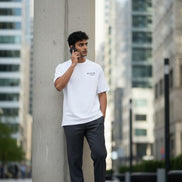 Man in a Billionaire by Orvella white t-shirt and dark pants leaning against a concrete pillar in an urban setting.