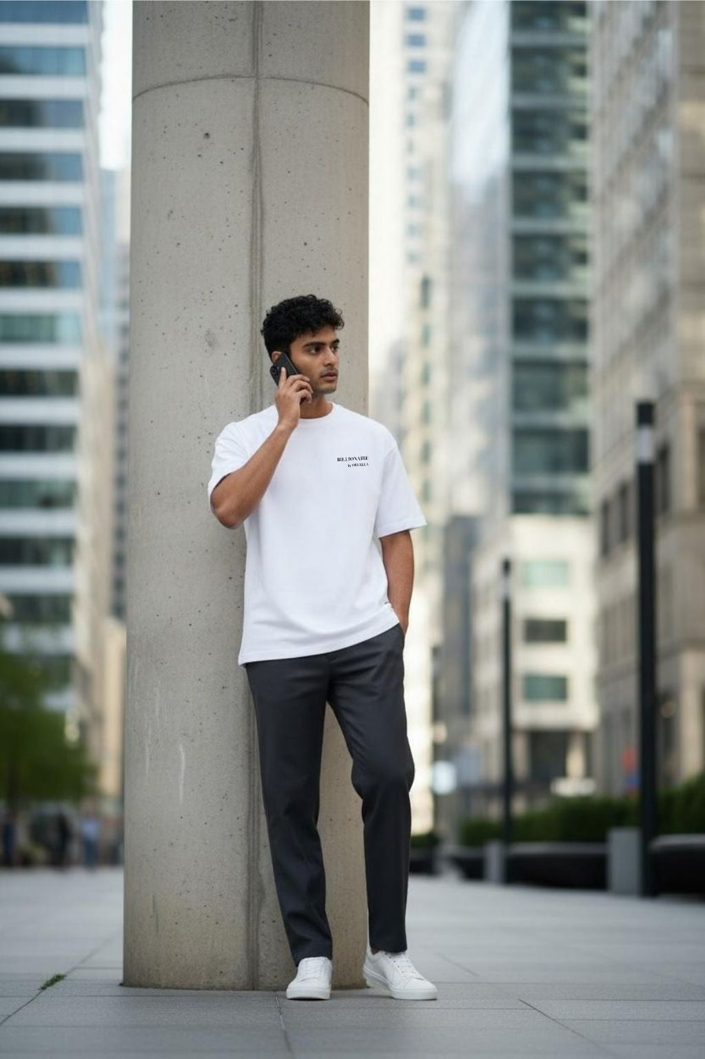 Man in a Billionaire by Orvella white t-shirt and dark pants leaning against a concrete pillar in an urban setting.