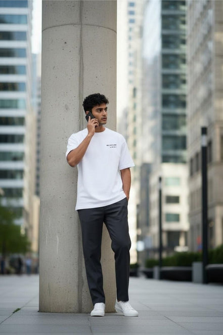 Man in a Billionaire by Orvella white t-shirt and dark pants leaning against a concrete pillar in an urban setting.