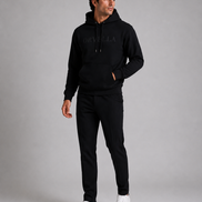 ORVELLA® Signature Hoodie in Jet Black worn by a male model in a luxury studio photoshoot. Premium heavyweight black hoodie with tonal ORVELLA branding and tonal sleeve logo, styled with black tailored trousers and white leather sneakers to showcase modern British luxury streetwear.