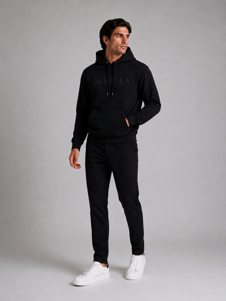 ORVELLA® Signature Hoodie in Jet Black worn by a male model in a luxury studio photoshoot. Premium heavyweight black hoodie with tonal ORVELLA branding and tonal sleeve logo, styled with black tailored trousers and white leather sneakers to showcase modern British luxury streetwear.