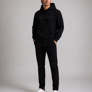 ORVELLA® Signature Hoodie in Jet Black worn by a male model in a professional studio photoshoot. Premium heavyweight black hoodie with tonal ORVELLA branding, paired with black tailored trousers and white leather sneakers, showcasing modern British luxury streetwear.