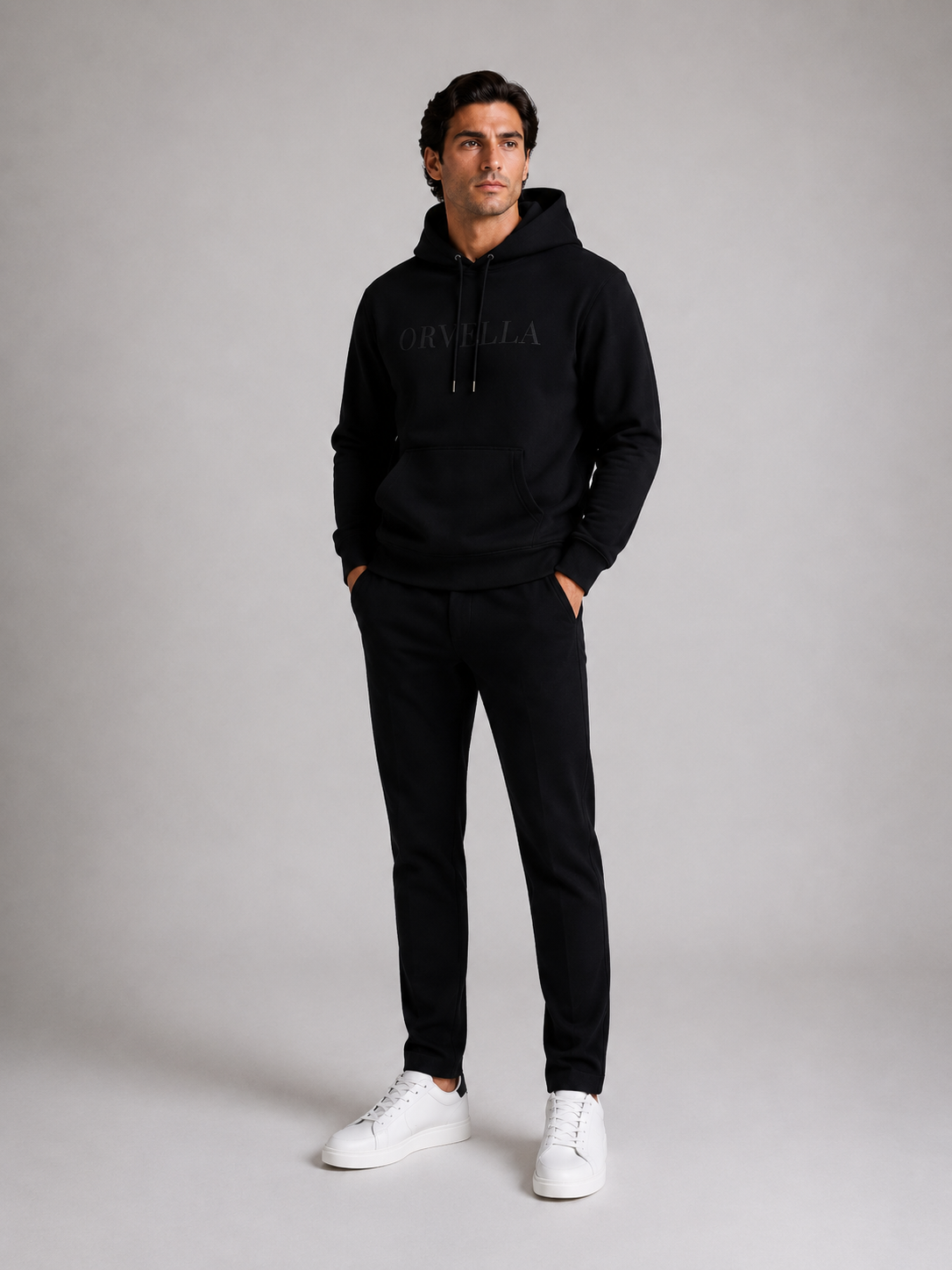 ORVELLA® Signature Hoodie in Jet Black worn by a male model in a professional studio photoshoot. Premium heavyweight black hoodie with tonal ORVELLA branding, paired with black tailored trousers and white leather sneakers, showcasing modern British luxury streetwear.