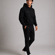 ORVELLA® Signature Hoodie in Jet Black worn by a male model in a luxury studio photoshoot. Premium heavyweight black hoodie with tonal ORVELLA branding and tonal sleeve logo, styled with black tailored trousers and white leather sneakers to showcase modern British luxury streetwear in a walking pose.