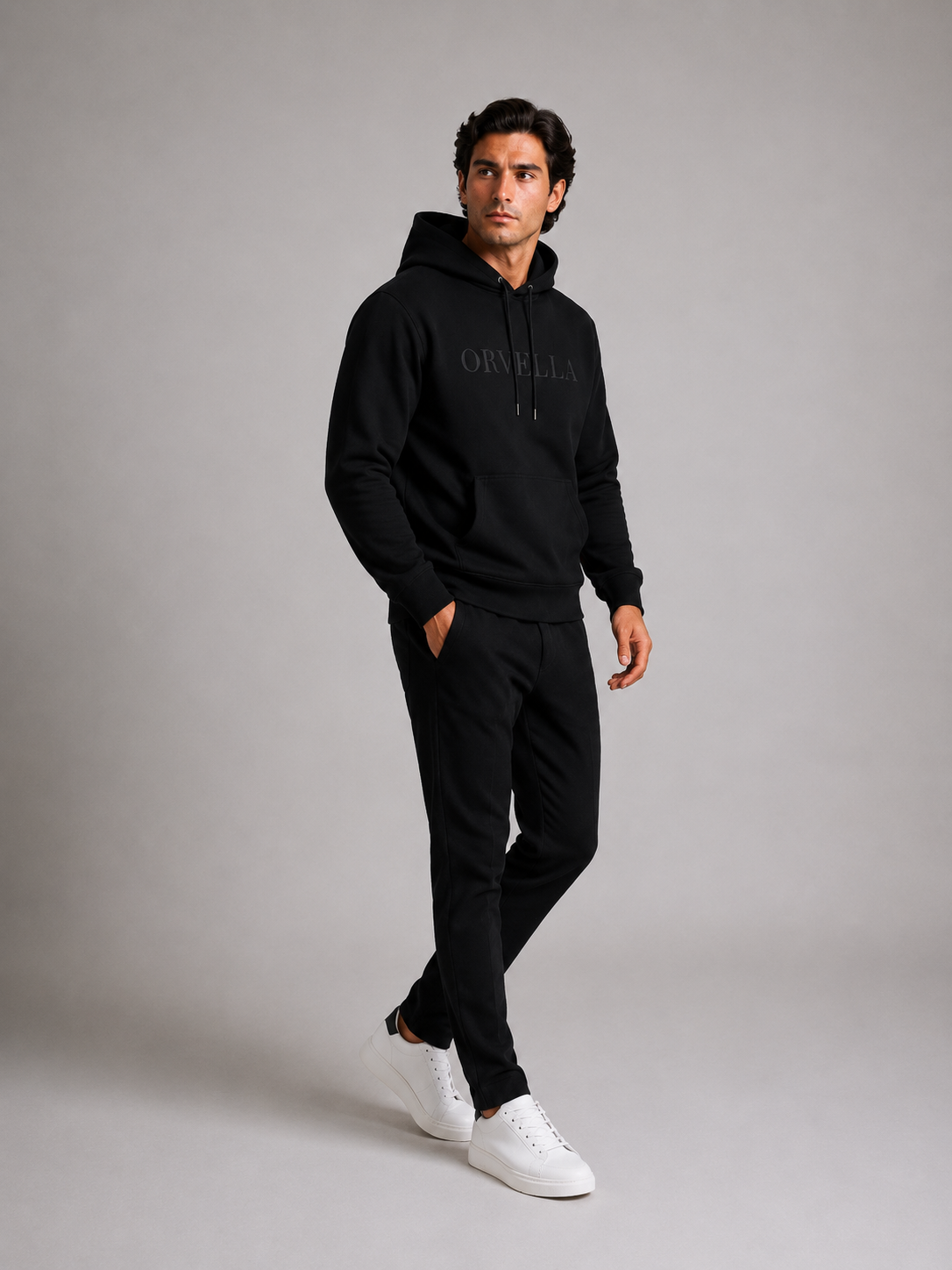 ORVELLA® Signature Hoodie in Jet Black worn by a male model in a luxury studio photoshoot. Premium heavyweight black hoodie with tonal ORVELLA branding and tonal sleeve logo, styled with black tailored trousers and white leather sneakers to showcase modern British luxury streetwear in a walking pose.