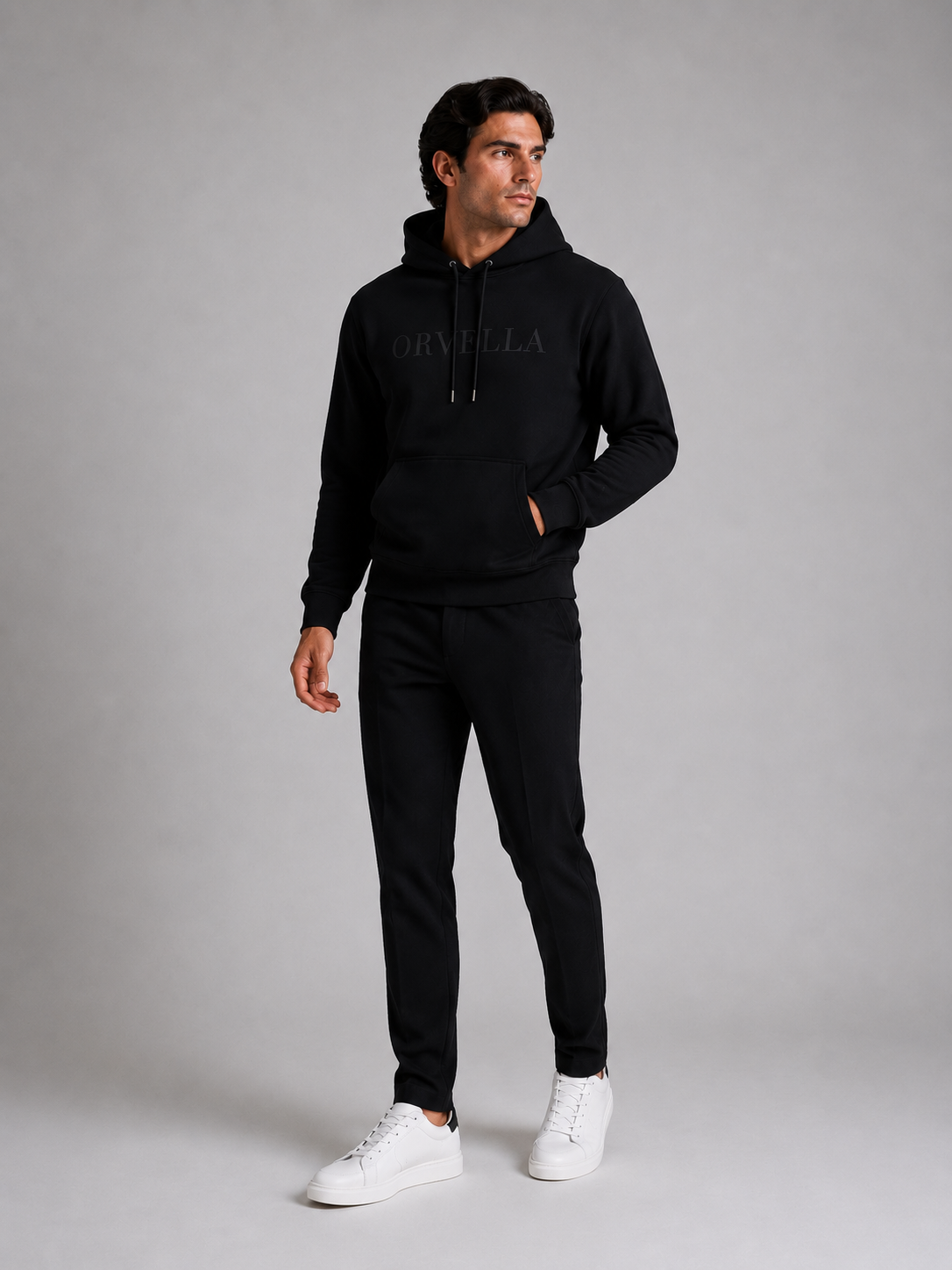 ORVELLA® Signature Hoodie in Jet Black worn by a male model in a luxury studio photoshoot. Premium heavyweight black hoodie with tonal ORVELLA branding and tonal sleeve logo, styled with black tailored trousers and white leather sneakers to showcase modern British luxury streetwear.