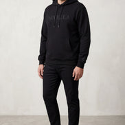 Male model wearing black ORVELLA luxury hoodie with tonal logo, black trousers and white trainers