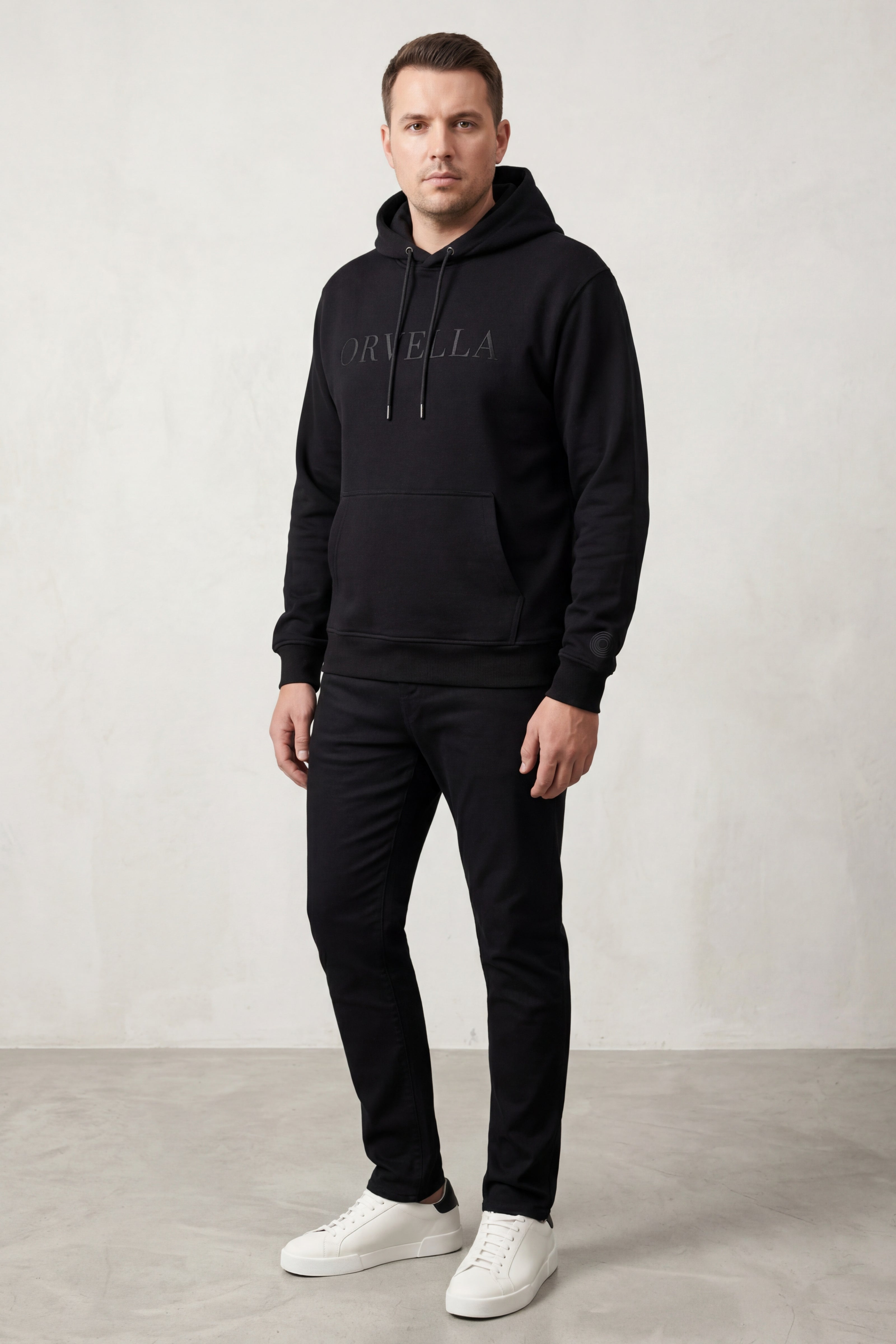 Male model wearing black ORVELLA luxury hoodie with tonal logo, black trousers and white trainers