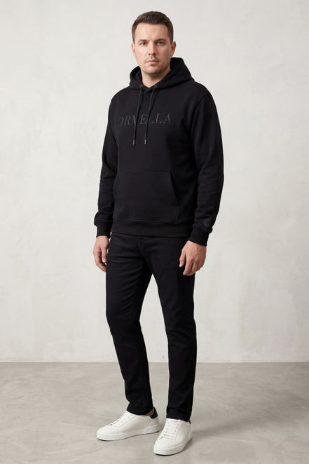 Male model wearing black ORVELLA luxury hoodie with tonal logo, black trousers and white trainers