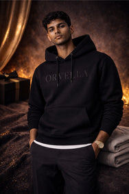Model wearing ORVELLA signature black hoodie with embossed logo, matte black aglets and premium fabric in a luxury studio setting.