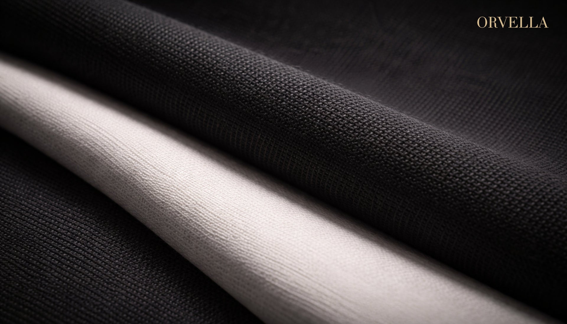 Close-up of layered black and white premium fabric with visible texture, softly lit in a luxury editorial style, featuring ORVELLA branding in the top corner.