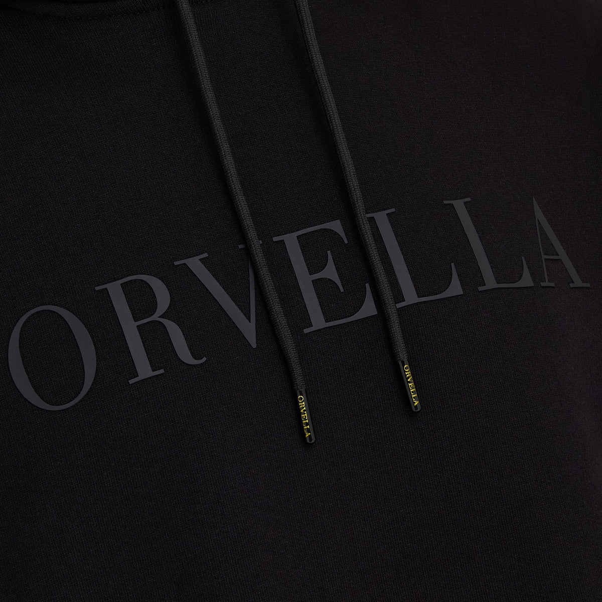 Close-up of the black ORVELLA luxury hoodie showing tonal chest branding, textured fabric, drawstrings, and branded aglet details.