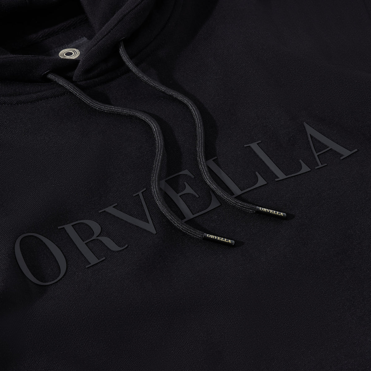 Close-up of the black ORVELLA luxury hoodie showing tonal chest branding, textured heavyweight fabric, drawstring hood, and branded aglet details.
