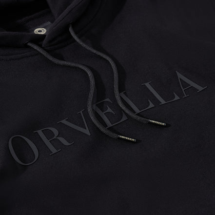 Close-up of the black ORVELLA luxury hoodie showing tonal chest branding, textured heavyweight fabric, drawstring hood, and branded aglet details.