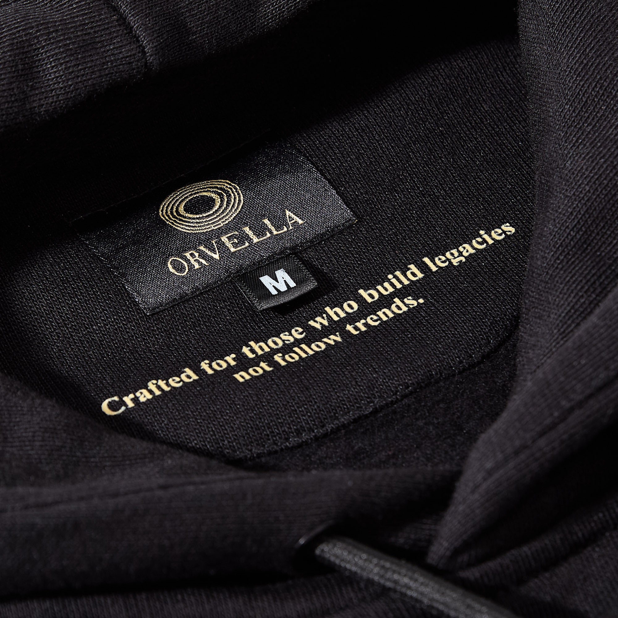 Close-up of the black ORVELLA luxury hoodie neck label, showing gold branding, size detail, premium fabric texture, and the inner quote “Crafted for those who build legacies not follow trends.”