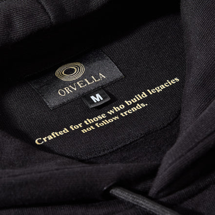 Close-up of the black ORVELLA luxury hoodie neck label, showing gold branding, size detail, premium fabric texture, and the inner quote “Crafted for those who build legacies not follow trends.”