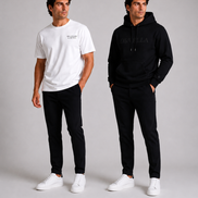 Two male models wearing ORVELLA menswear against a light grey studio background: one in a white Billionaire by ORVELLA T-shirt and black trousers, the other in a black ORVELLA hoodie and matching trousers.