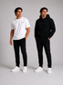 Two male models wearing ORVELLA menswear against a light grey studio background: one in a white Billionaire by ORVELLA T-shirt and black trousers, the other in a black ORVELLA hoodie and matching trousers.