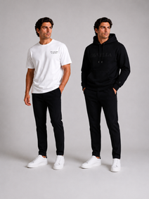 Two male models wearing ORVELLA menswear against a light grey studio background: one in a white Billionaire by ORVELLA T-shirt and black trousers, the other in a black ORVELLA hoodie and matching trousers.